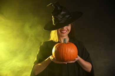 Photo of Young woman wearing witch costume with pumpkin in smoke cloud on dark background. Halloween party