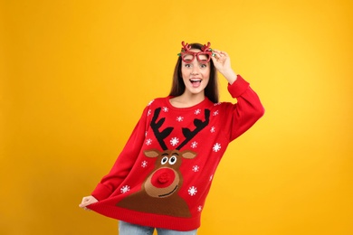 Surprised young woman in Christmas sweater and party glasses on yellow background