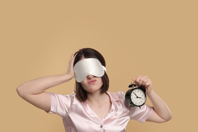 Tired young woman with sleep mask and alarm clock on beige background. Insomnia problem