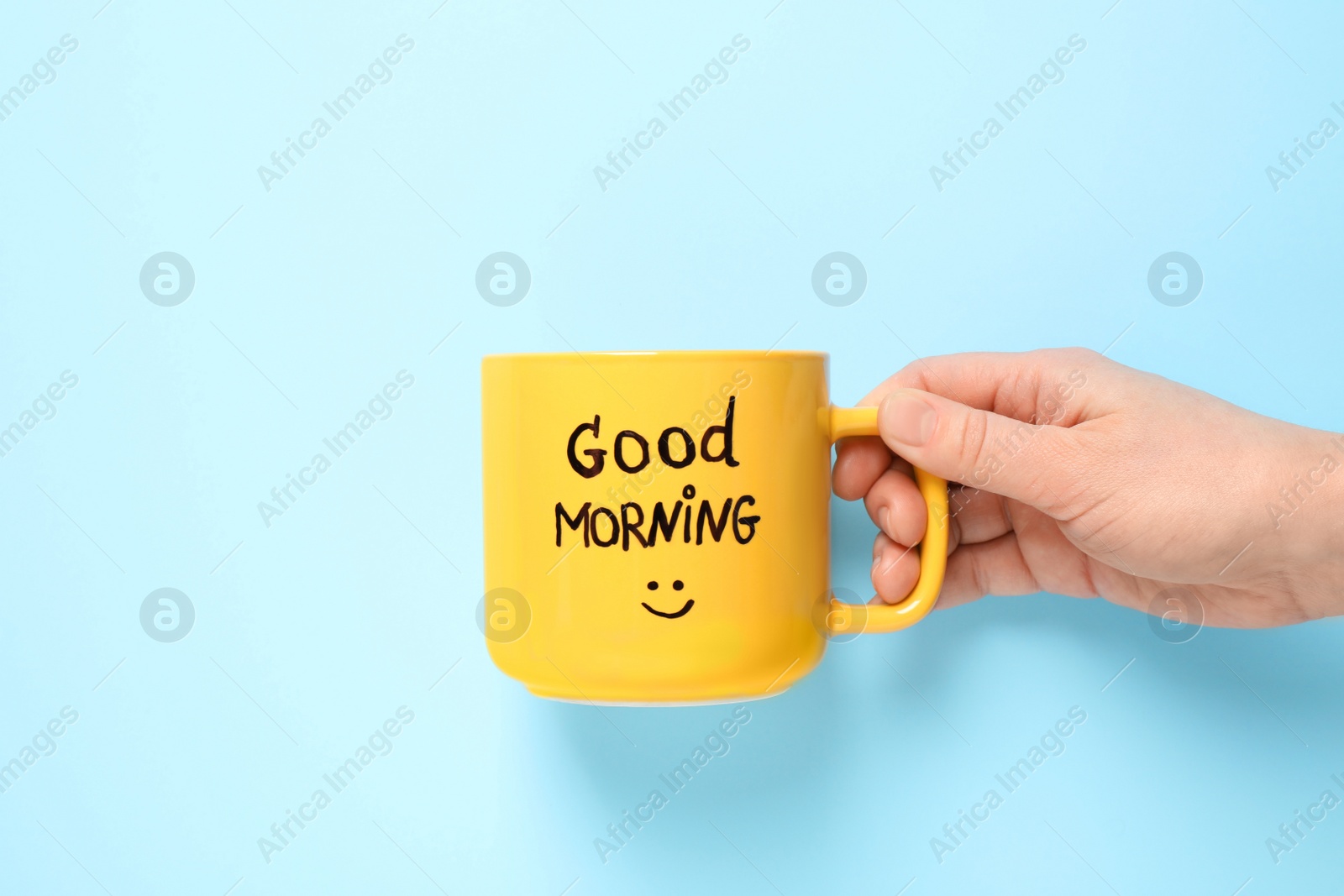 Photo of Woman holding cup of coffee with words GOOD MORNING on light blue background, closeup