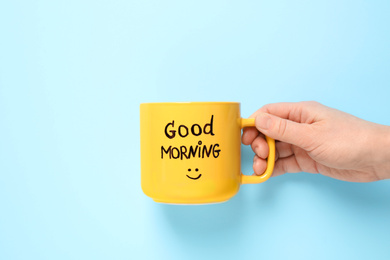 Woman holding cup of coffee with words GOOD MORNING on light blue background, closeup