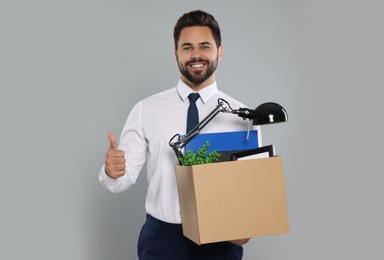 Photo of Happy unemployed man with box of personal office belongings showing thumb up on light grey background