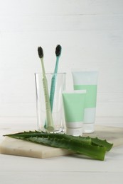 Tubes of toothpaste, toothbrushes and fresh aloe vera leaves on white wooden table