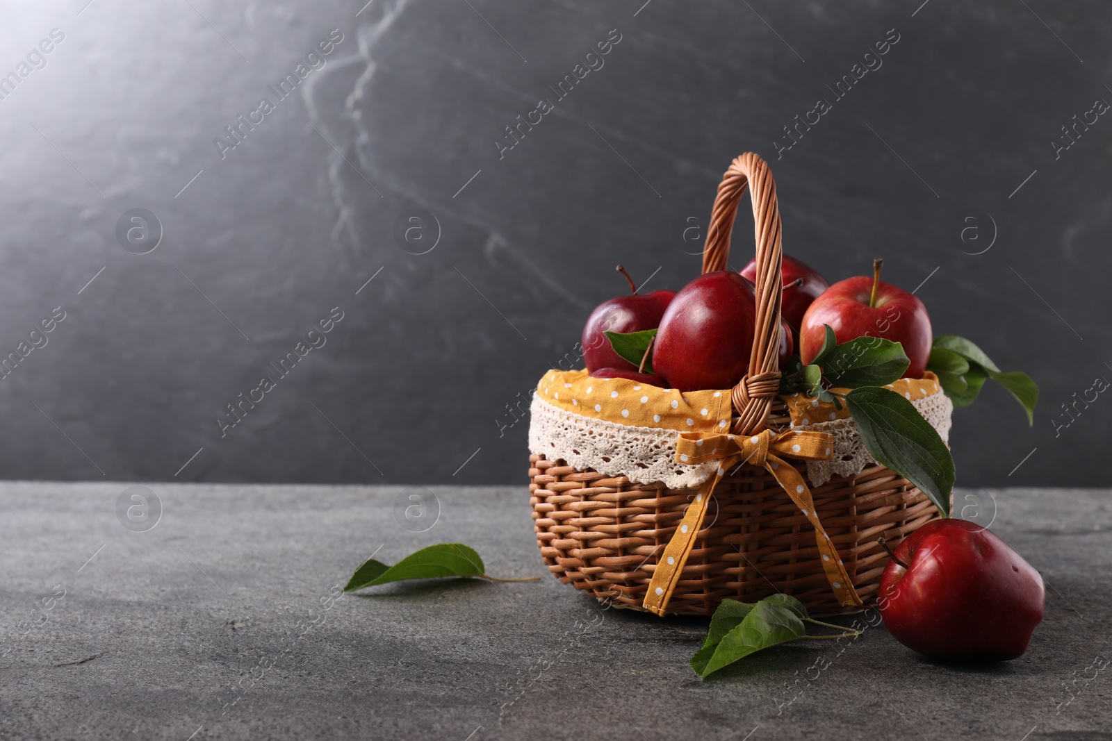 Photo of Ripe red apples and leaves in wicker basket on dark grey table. Space for text