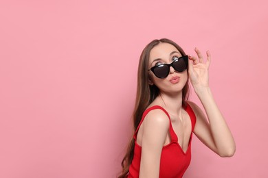 Photo of Beautiful young woman with sunglasses blowing kiss on pink background, space for text