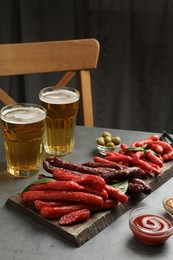 Photo of Different thin dry smoked sausages, sauces and glasses of beer on grey table