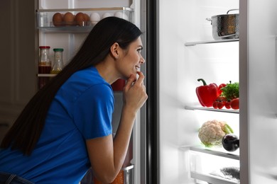Photo of Thoughtful young woman looking into modern refrigerator at night