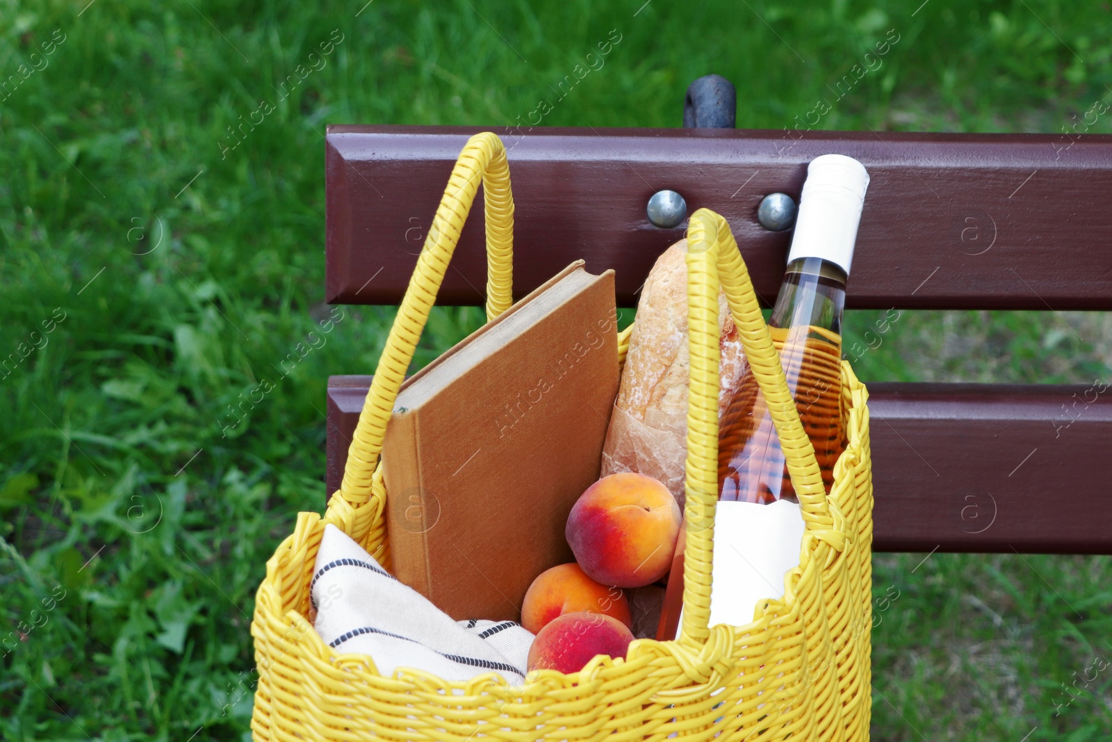 Photo of Yellow wicker bag with book, peaches, baguette and wine on wooden bench outdoors