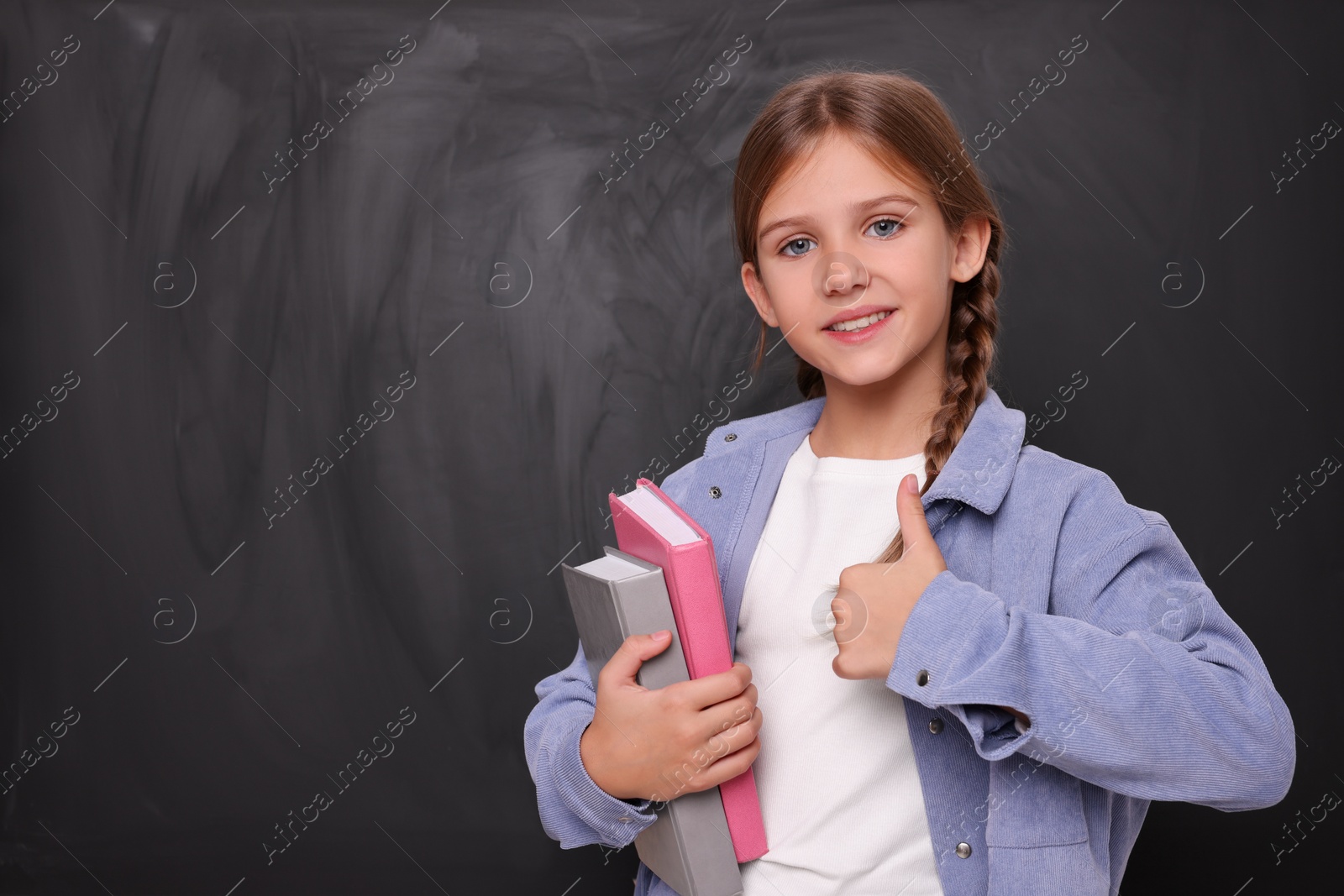 Photo of Smiling schoolgirl with books showing thumb up near blackboard. Space for text