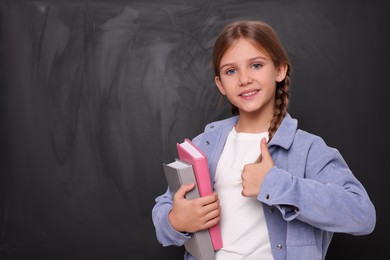 Photo of Smiling schoolgirl with books showing thumb up near blackboard. Space for text
