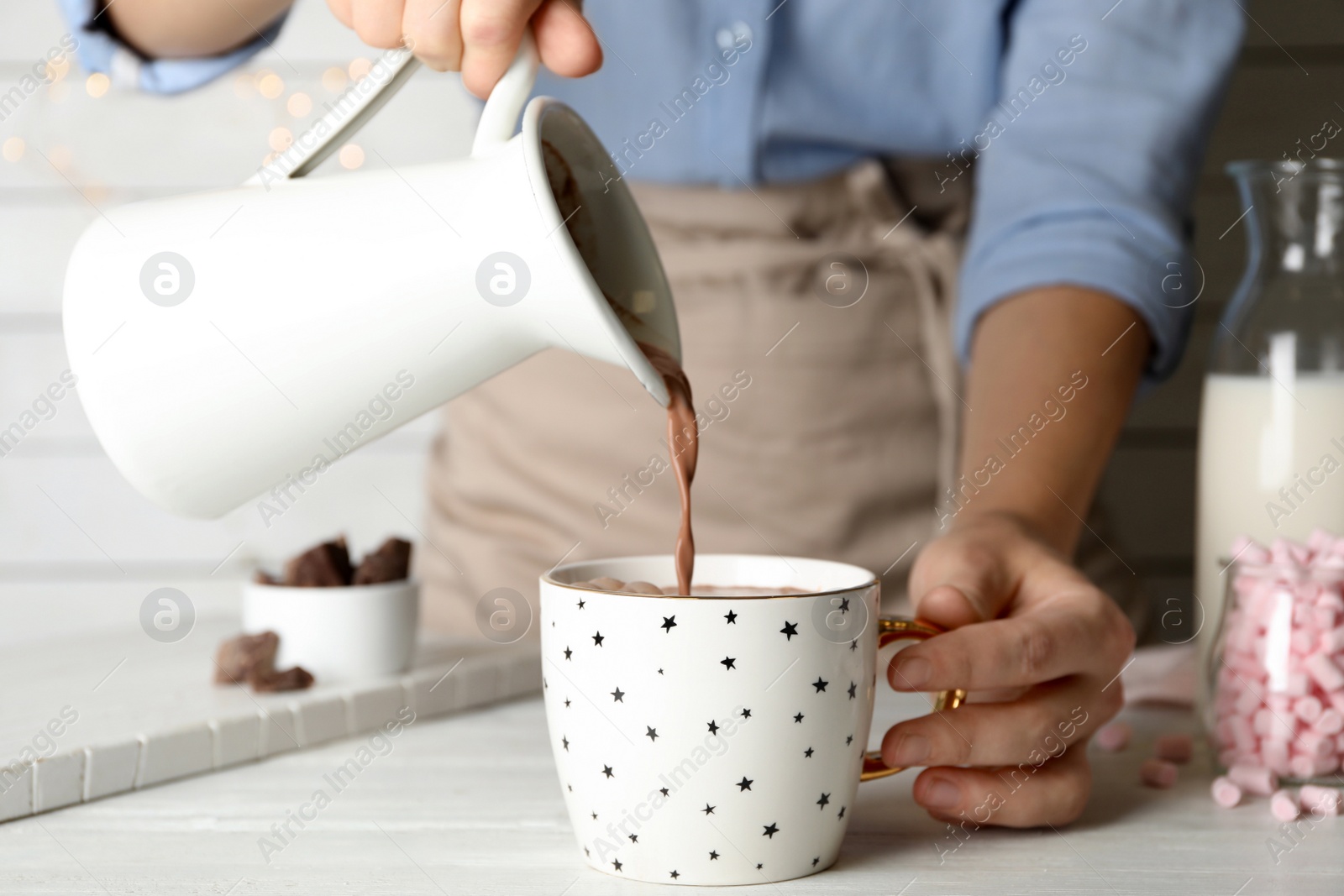 Photo of Woman pouring hot cocoa drink into cup on white wooden table, closeup