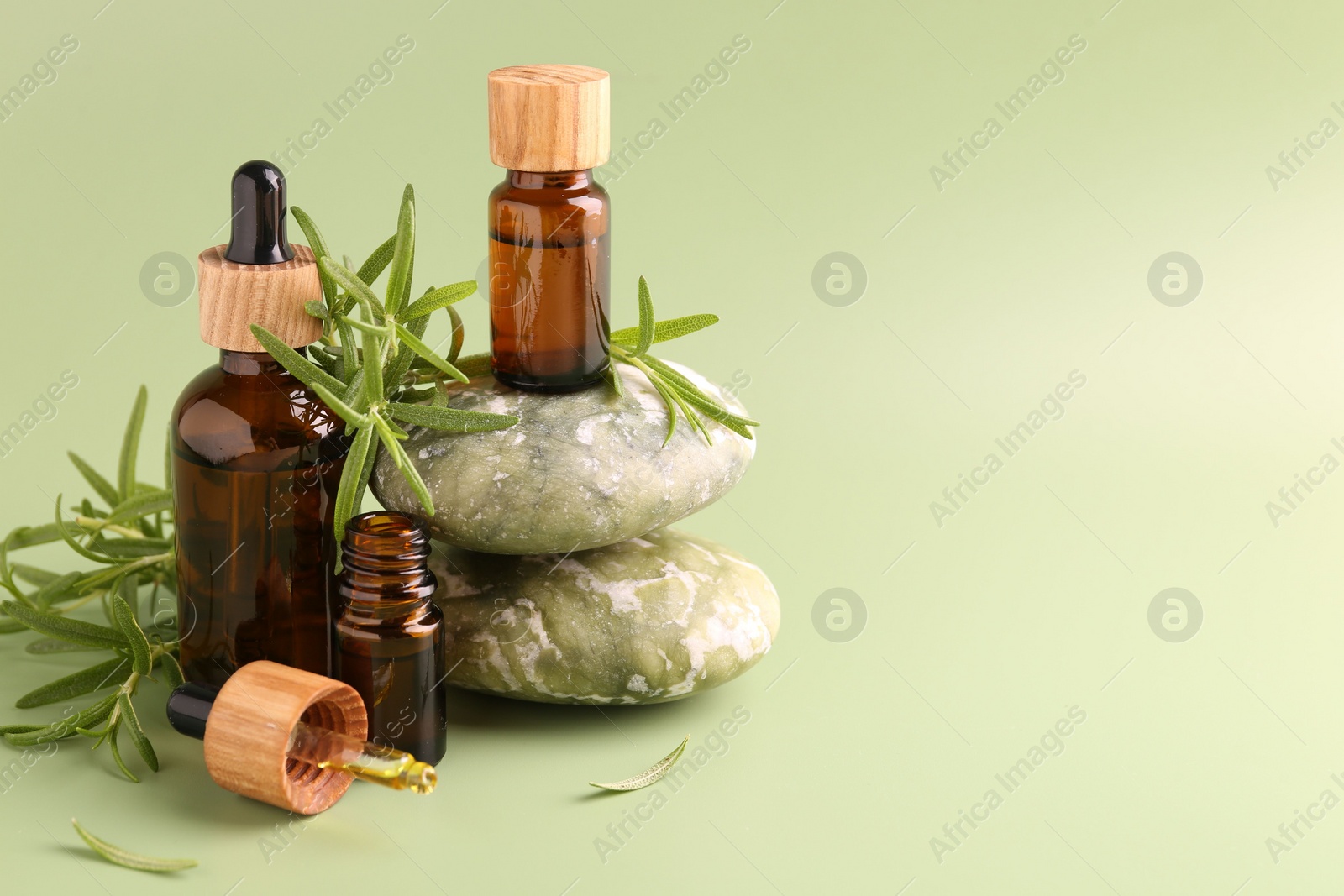 Photo of Aromatic essential oils in bottles, rosemary, pipette and pebble stones on green background. Space for text