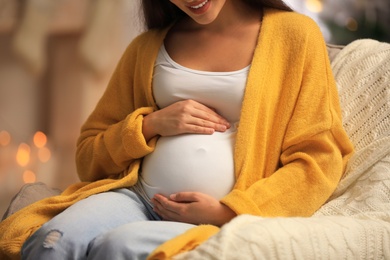 Happy pregnant woman in armchair at room decorated for Christmas, closeup. Expecting baby