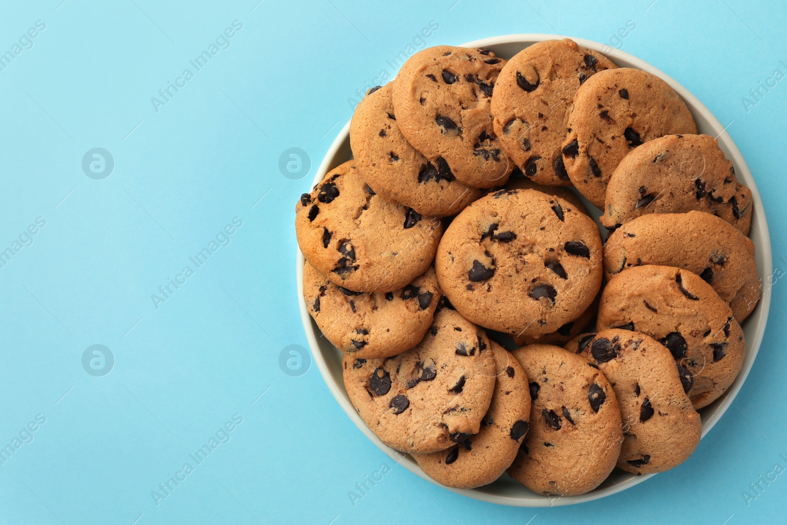 Photo of Bowl of delicious chocolate chip cookies on light blue background, top view. Space for text