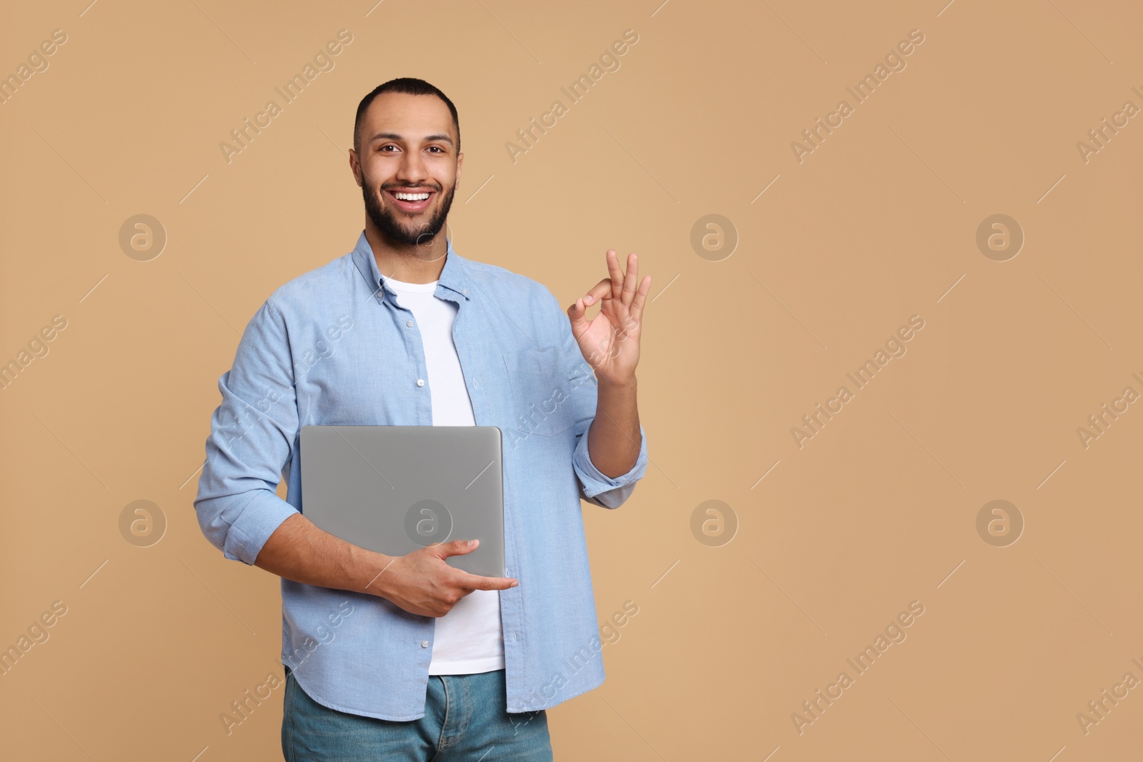 Photo of Smiling young man with laptop showing OK gesture on beige background, space for text