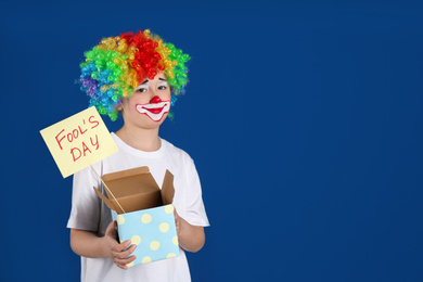 Photo of Preteen boy with clown makeup and wig holding sign APRIL FOOL'S DAY on blue background, space for text