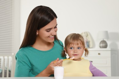 Mother feeding her cute little child with yogurt at white table in room