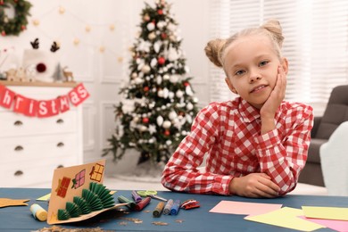 Photo of Cute little child with materials and beautiful Christmas card at table in decorated room