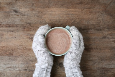 Woman with knitted mittens holding cup of delicious cocoa drink at wooden table, top view