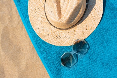 Soft blue beach towel with straw hat and sunglasses on sand, flat lay