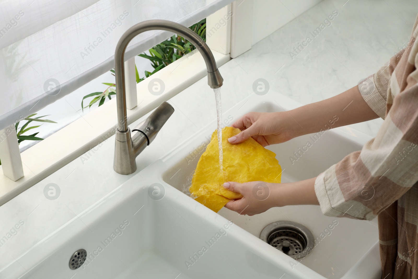 Photo of Woman washing beeswax food wrap under tap water in kitchen sink, closeup