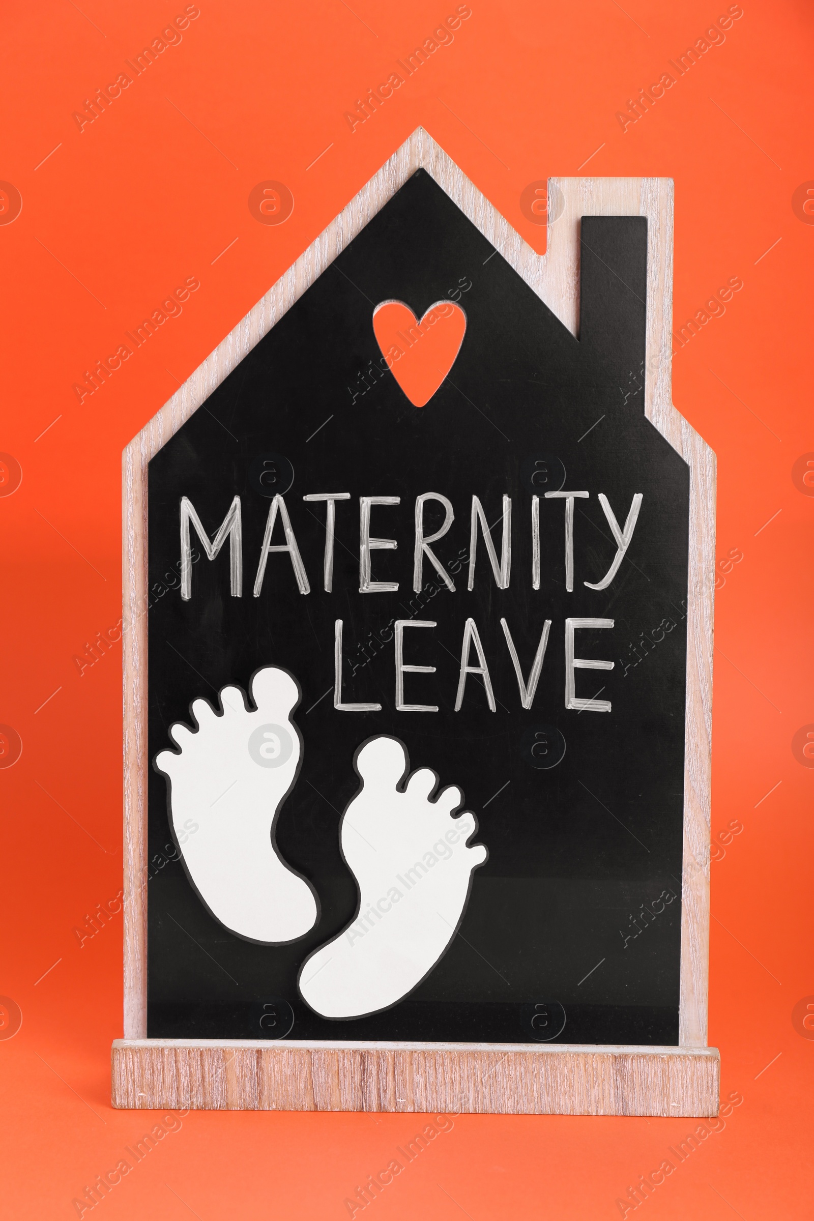 Photo of Wooden house figure with words Maternity Leave and paper cutout of baby feet on orange background