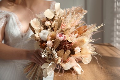 Photo of Bride holding beautiful dried flower bouquet near window at home, closeup