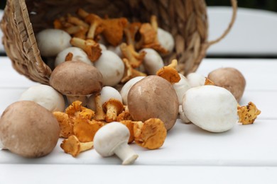 Photo of Overturned wicker basket with different fresh mushrooms on white wooden table, closeup