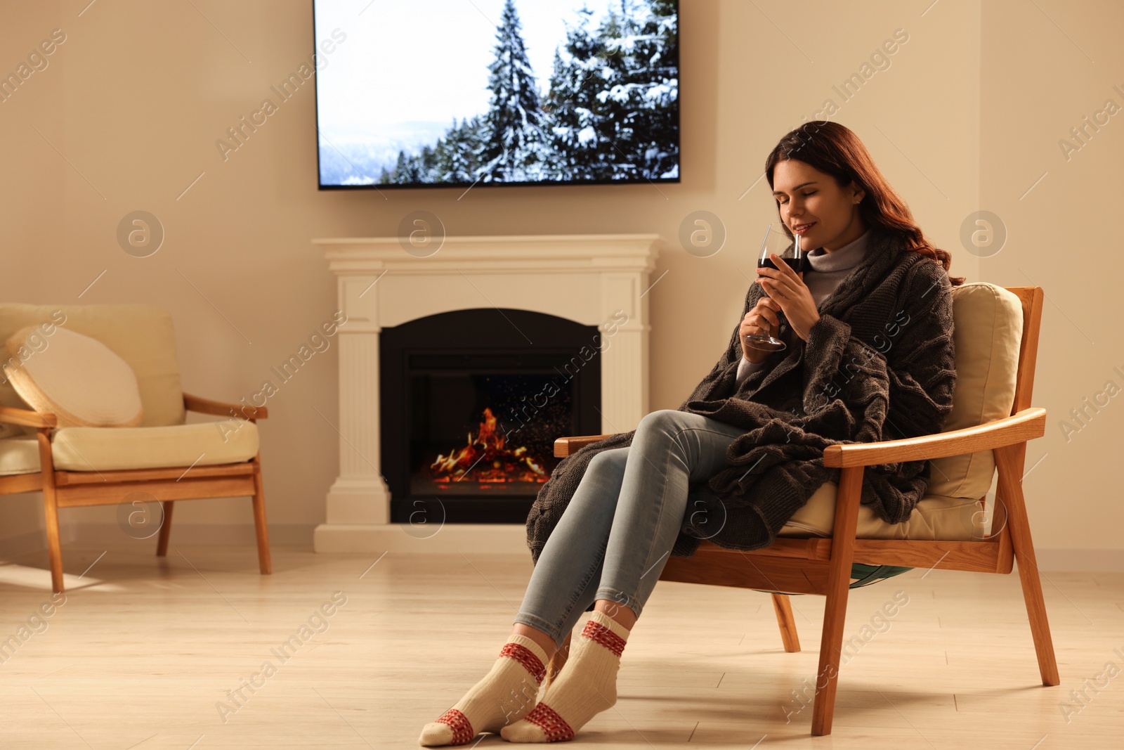 Photo of Young woman with glass of wine resting in armchair near fireplace at home