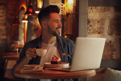Photo of Young blogger with laptop eating dessert and drinking coffee at table in cafe