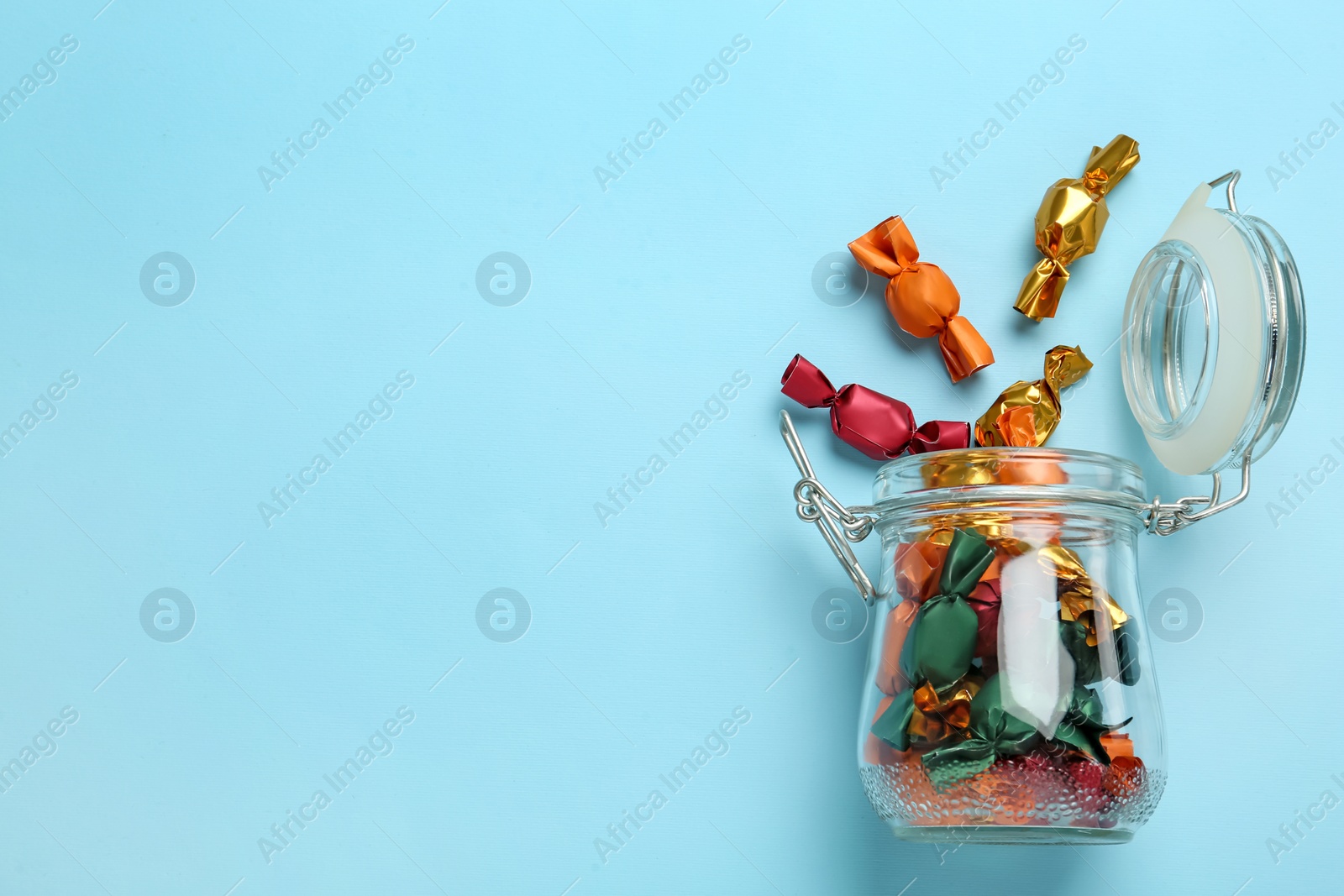 Photo of Overturned glass jar with candies in colorful wrappers on light blue background, top view. Space for text