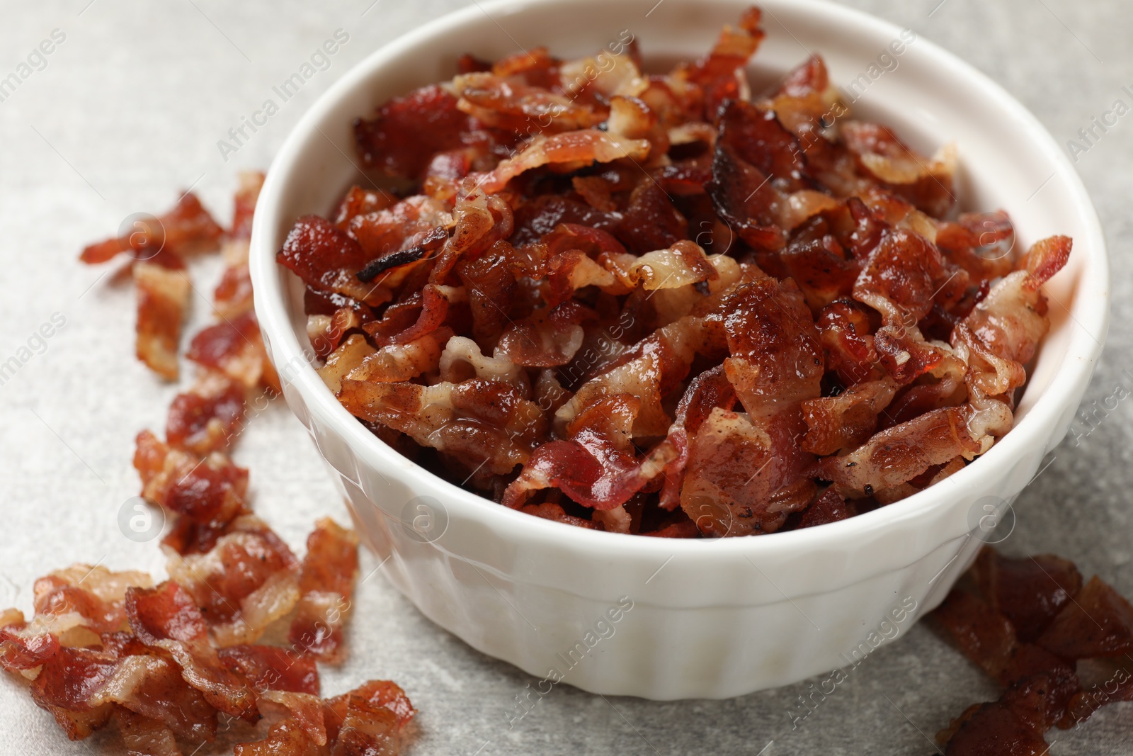 Photo of Pieces of tasty fried bacon in bowl on gray textured table, closeup