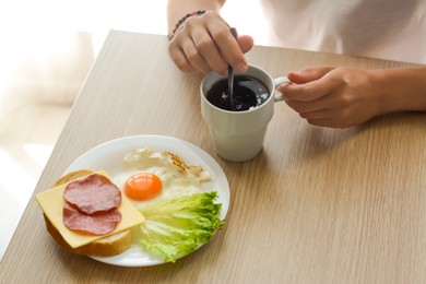 Woman stirring morning coffee while having breakfast at wooden table indoors, closeup