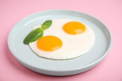 Tasty fried eggs with basil in plate on pink background, closeup