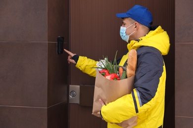 Photo of Courier in medical mask holding paper bag with groceries and ringing doorbell outdoors. Delivery service during quarantine due to Covid-19 outbreak