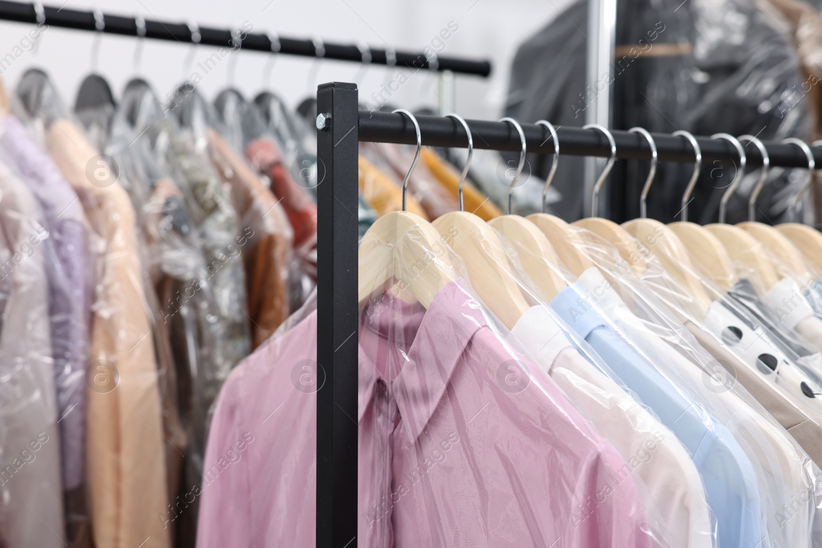 Photo of Dry-cleaning service. Many different clothes in plastic bags hanging on rack indoors, closeup