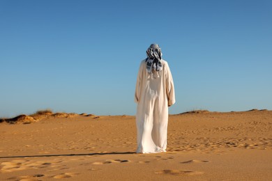 Man in arabic clothes walking through desert on sunny day, back view