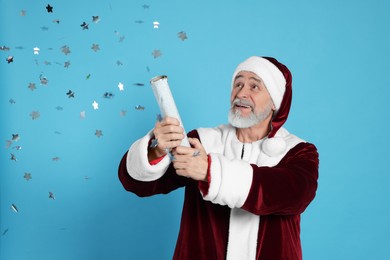 Man in Santa Claus costume blowing up party popper on light blue background