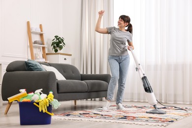 Happy young housewife vacuuming rug at home