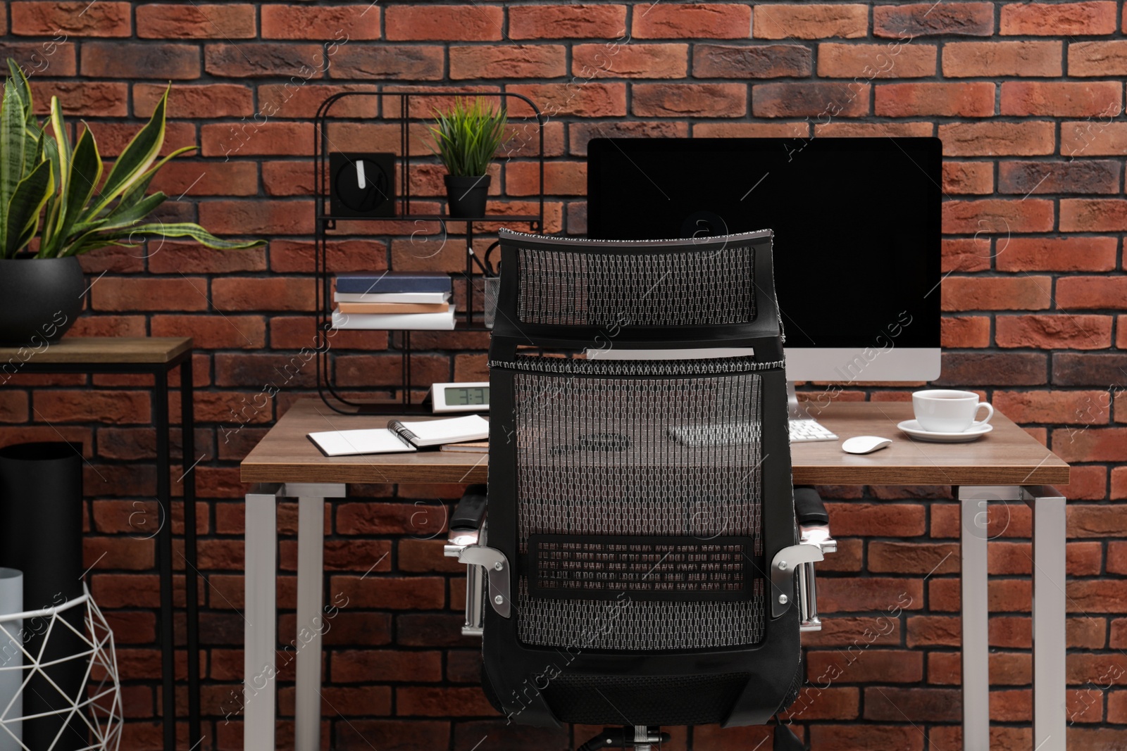 Photo of Cosy workspace with computer on desk, chair and potted plants near brick wall at home