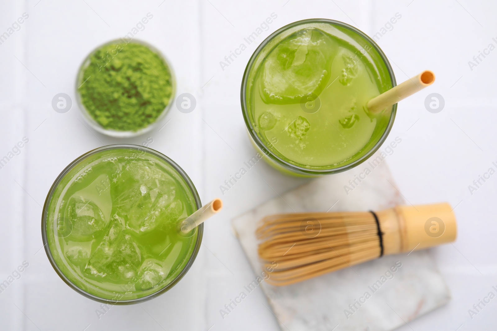 Photo of Delicious iced green matcha tea, powder and bamboo whisk on white tiled table, flat lay