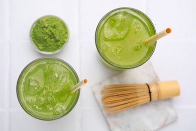 Delicious iced green matcha tea, powder and bamboo whisk on white tiled table, flat lay