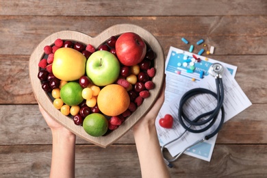 Photo of Female doctor holding plate with fresh fruits over wooden table, top view. Cardiac diet