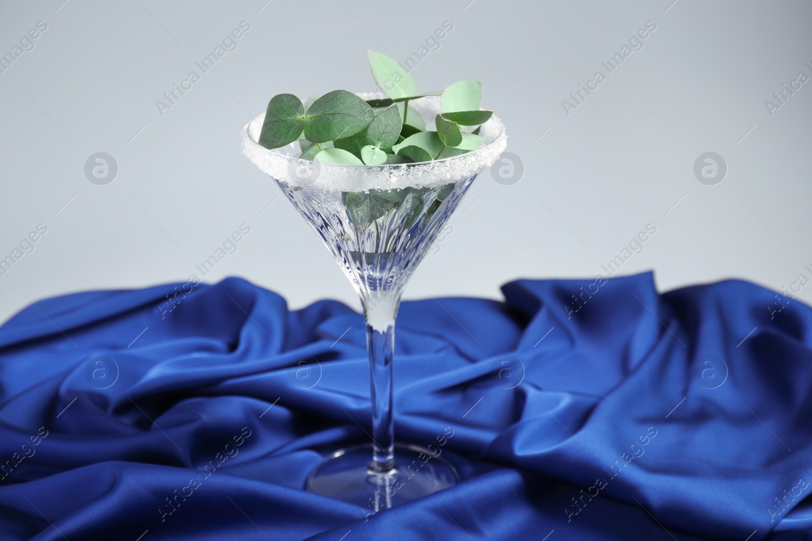 Photo of Beautiful martini glass with sugar rim and eucalyptus leaves on blue satin against light grey background