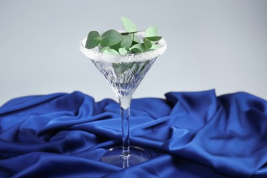 Photo of Beautiful martini glass with sugar rim and eucalyptus leaves on blue satin against light grey background