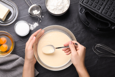 Woman preparing dough for Belgian waffles at black table, top view