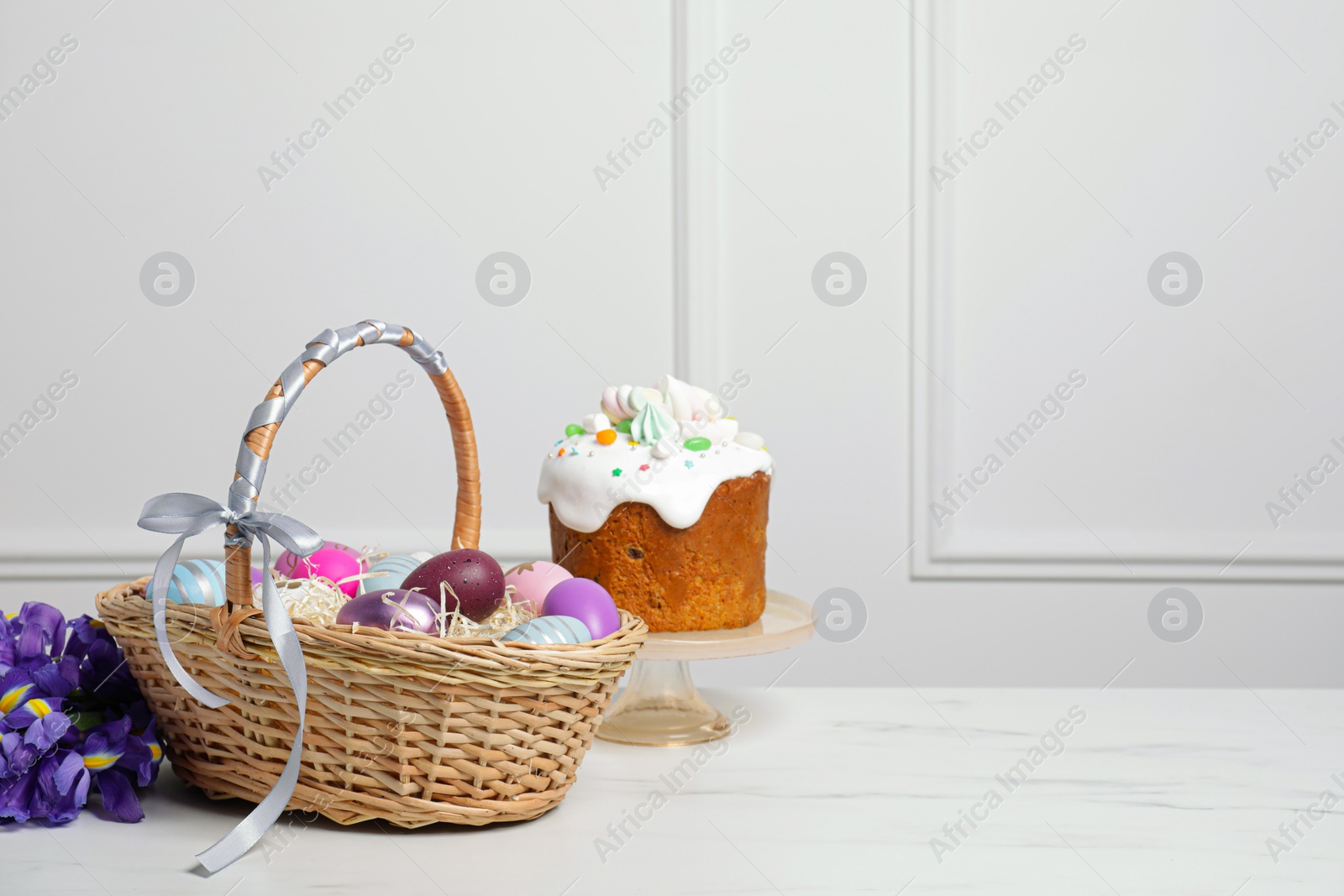 Photo of Easter basket with painted eggs near tasty cake and bouquet of flowers on white marble table. Space for text