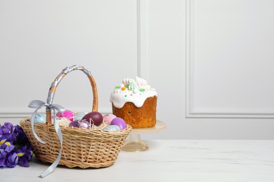 Photo of Easter basket with painted eggs near tasty cake and bouquet of flowers on white marble table. Space for text