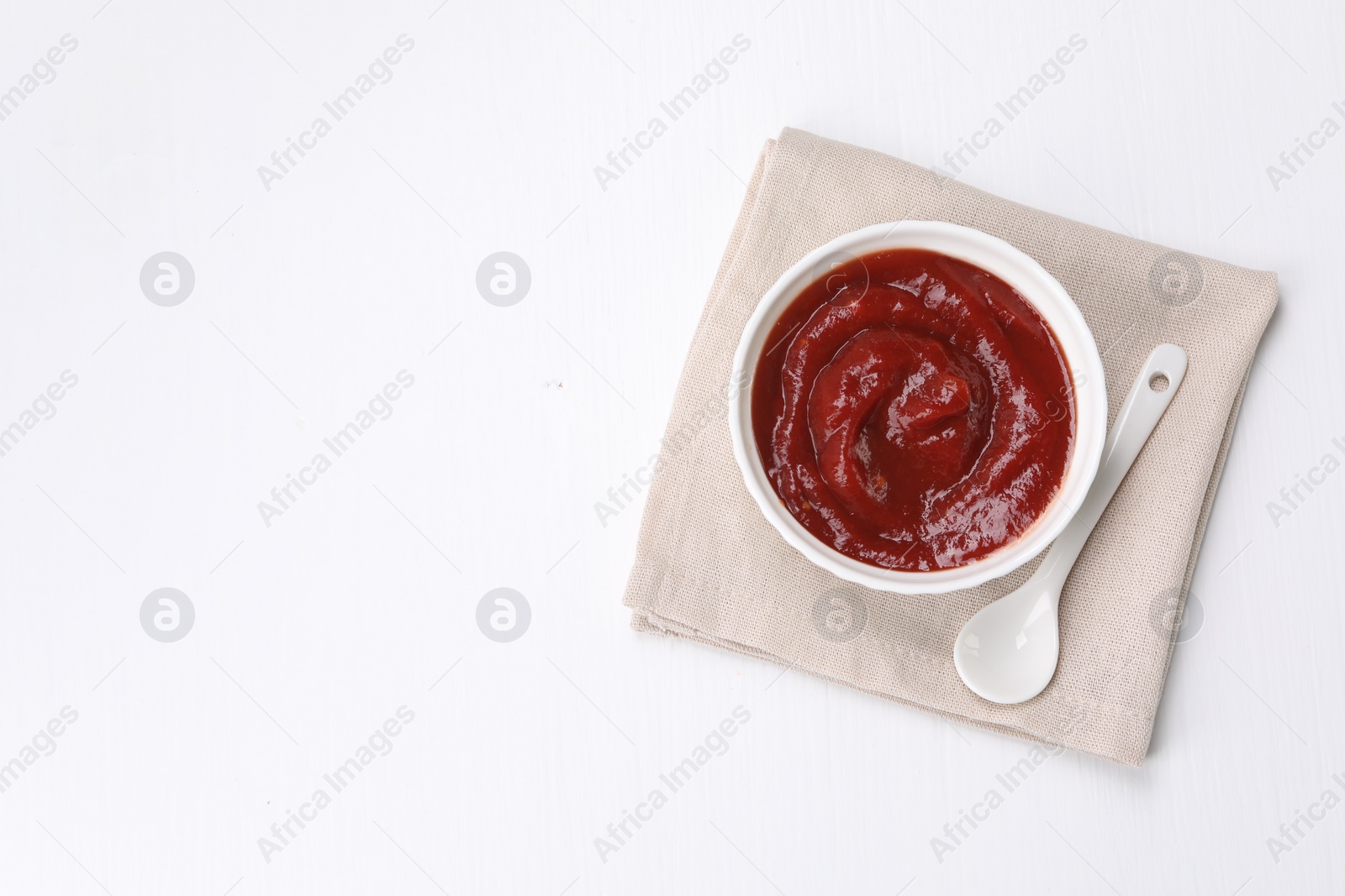 Photo of Organic ketchup in bowl and spoon on white table, top view with space for text. Tomato sauce