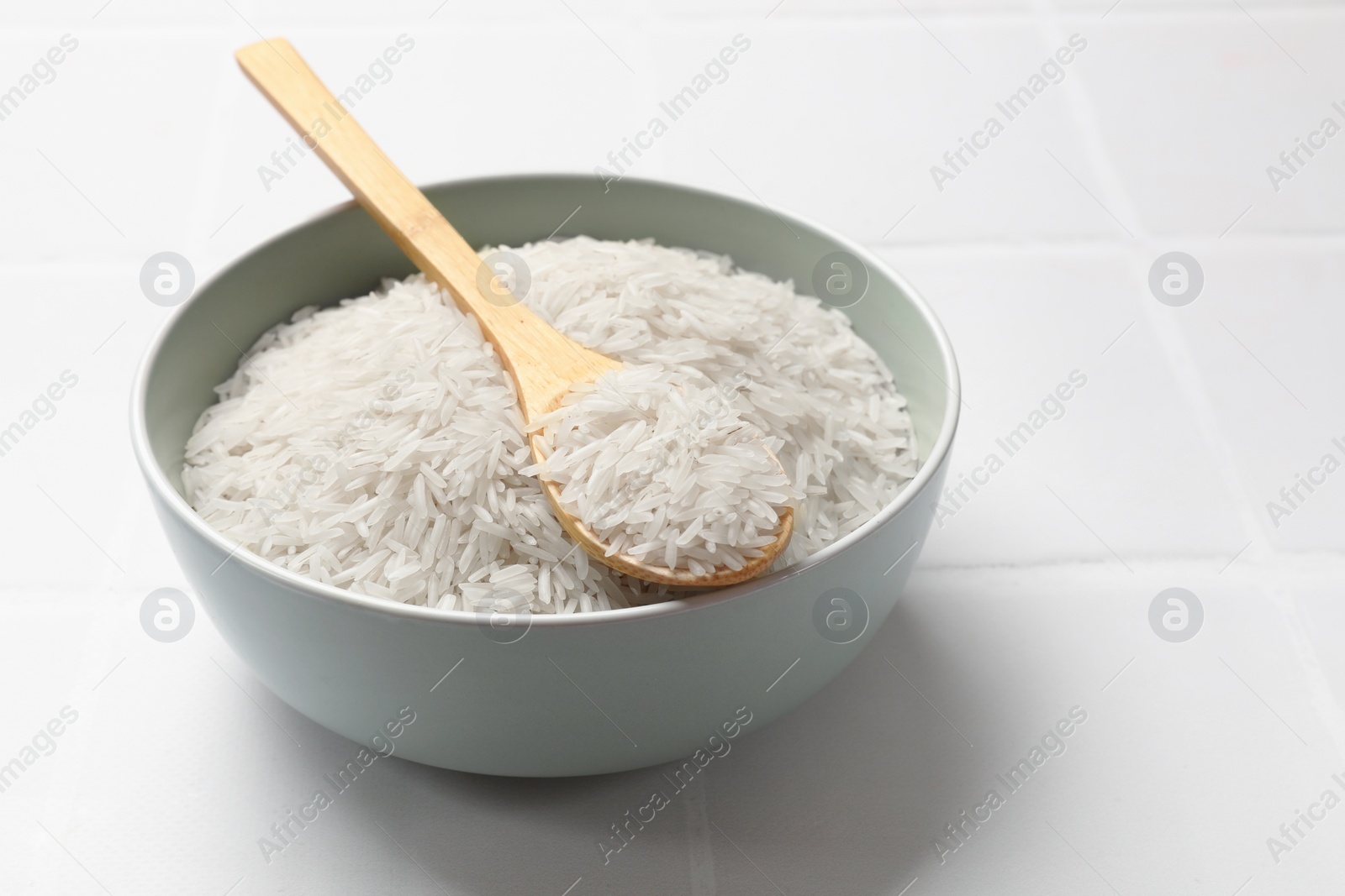 Photo of Raw basmati rice in bowl and spoon on white tiled table